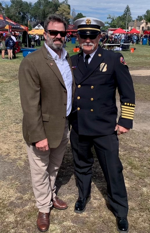 Two men smiling, one in a fire chief uniform, the other in a blazer, at an outdoor event.
