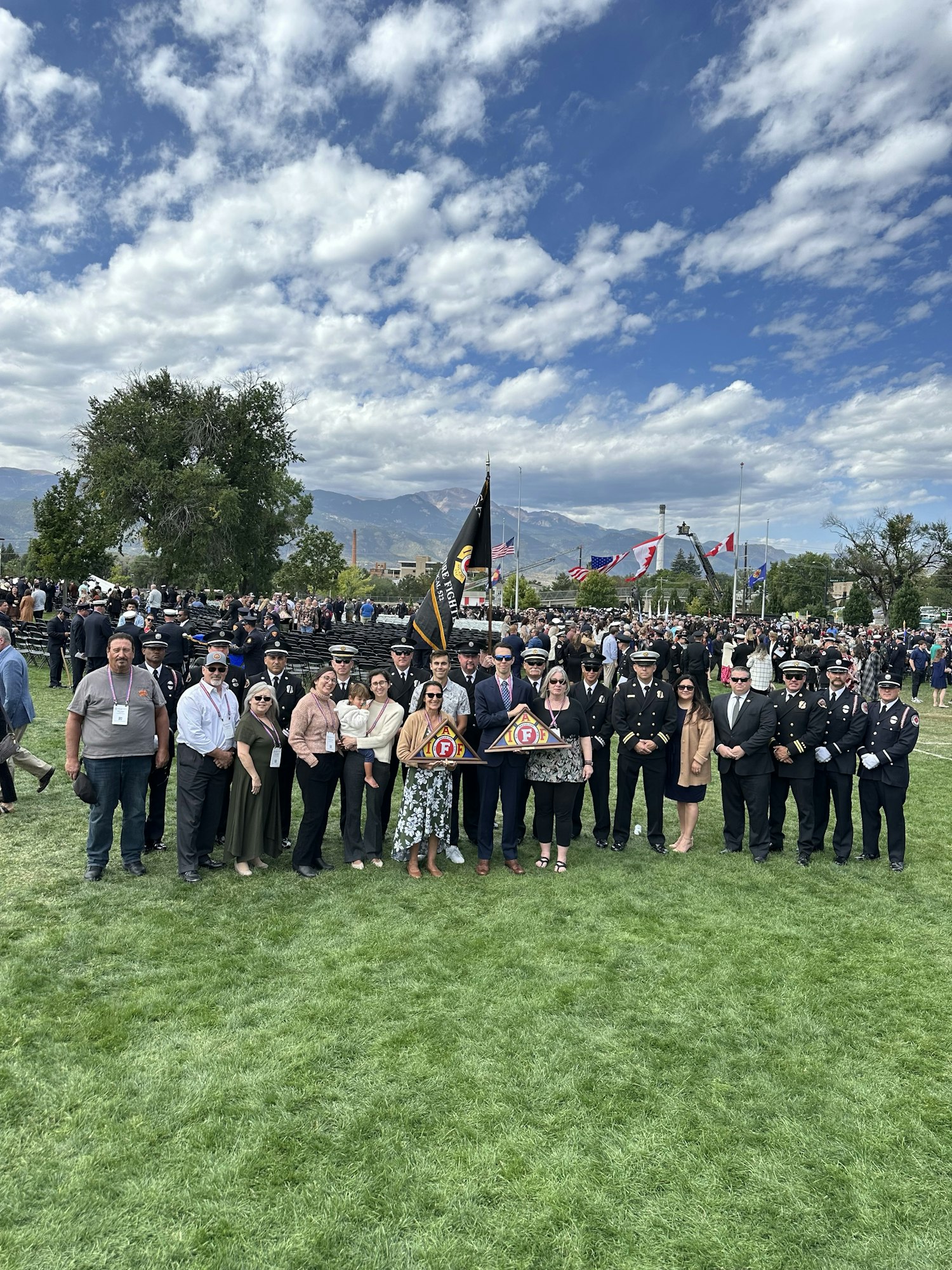 Group of people at an outdoor event with flags, uniforms suggesting a formal or commemorative occasion, under a cloudy sky.