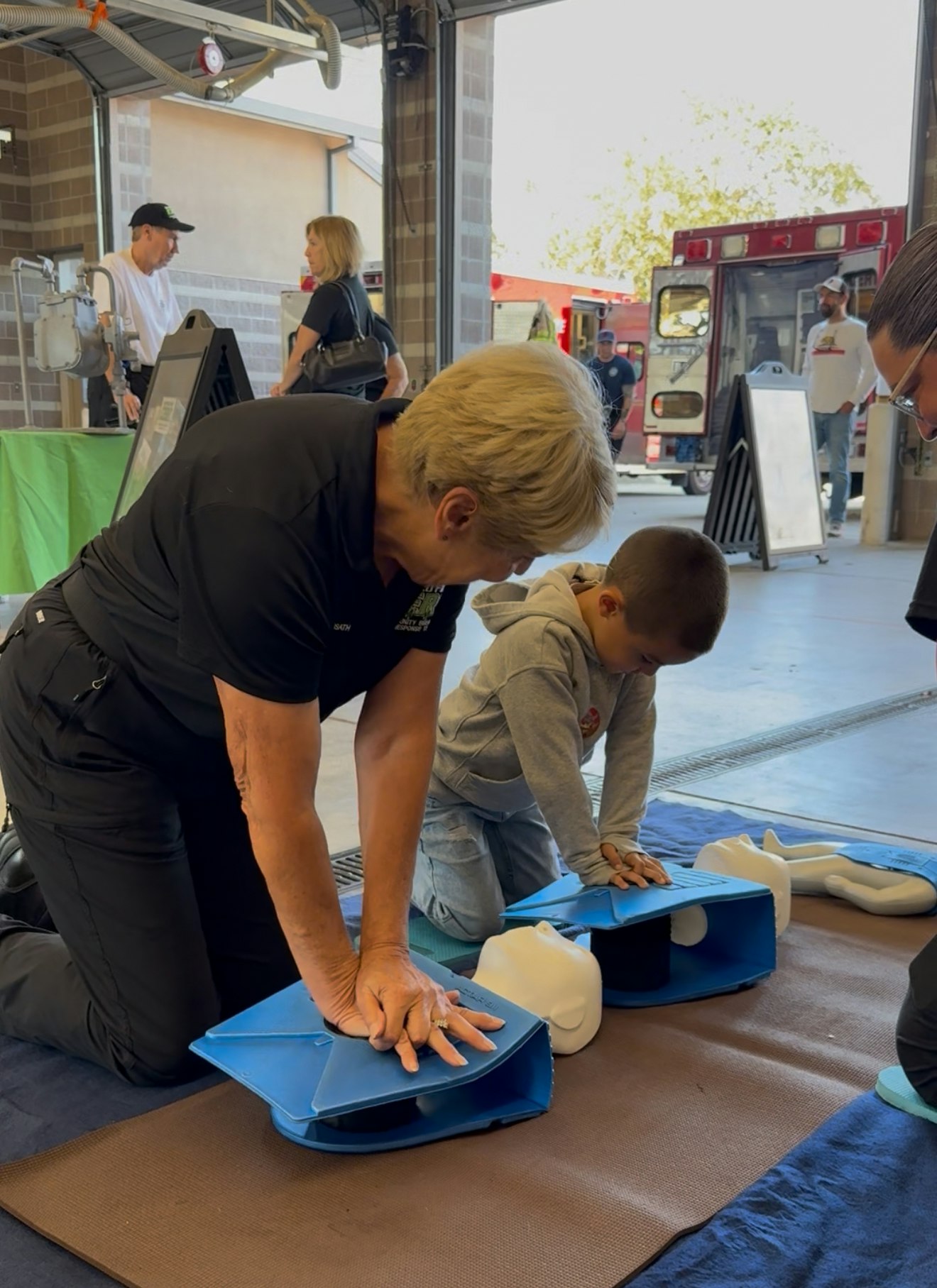 A woman and a young boy practice CPR on training mannequins in a fire station, with others present in the background.