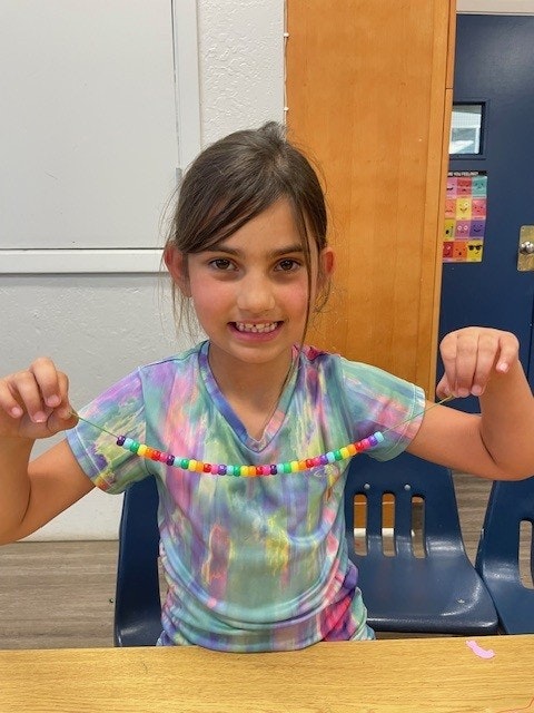 A child in a colorful shirt smiles while holding up a string of colorful beads, seated at a table indoors.