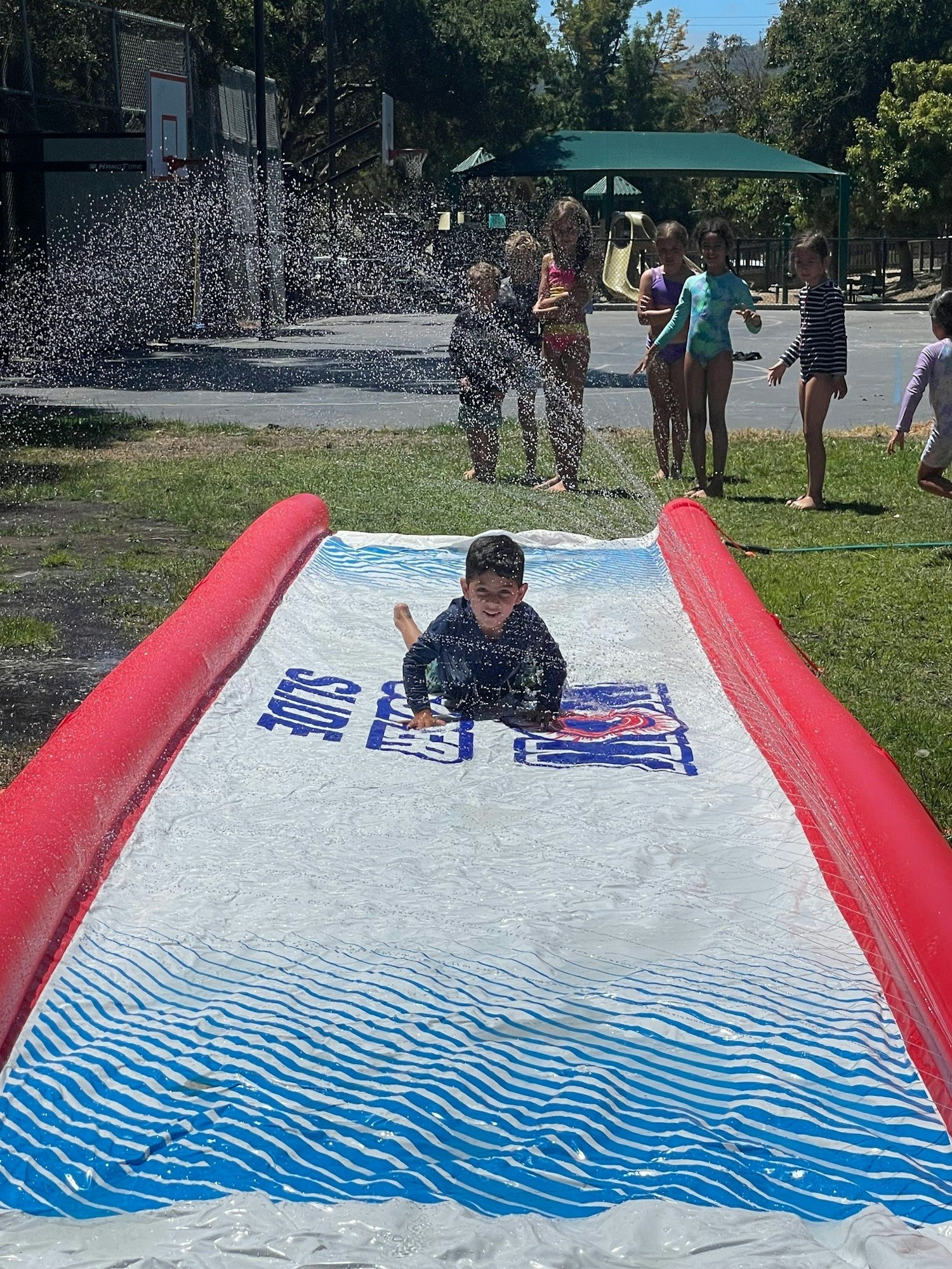 A child sliding on a water slide while others watch, with water splashing around, in a park setting.