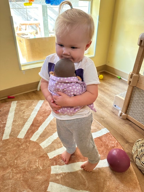 A toddler holding a doll, standing in a room with toys and a sun-patterned rug.