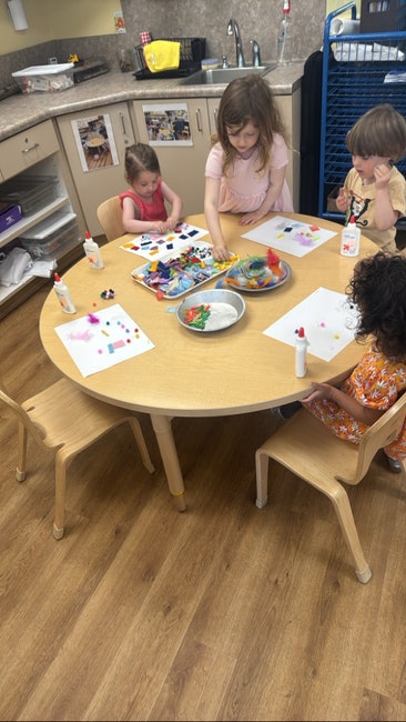 Children are making art using paper, glue, and colorful materials at a round table in a classroom.