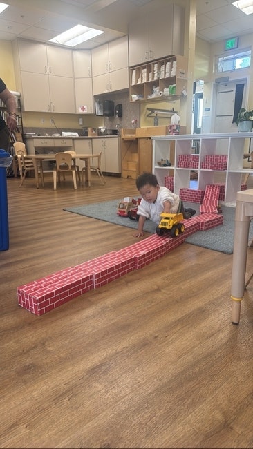 Child playing with toy trucks on brick-patterned blocks in a classroom.