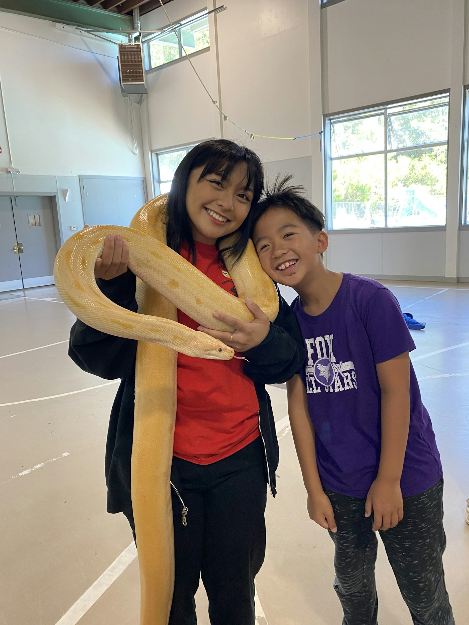 A girl holding a large yellow snake smiles with a boy beside her, both appearing happy in a spacious indoor setting.