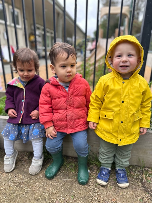 Three toddlers in colorful jackets sitting in front of a fence, with a playground in the background.