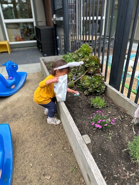 A child in a yellow shirt collects Easter eggs in a garden with flowers and plants, holding a paper bag.