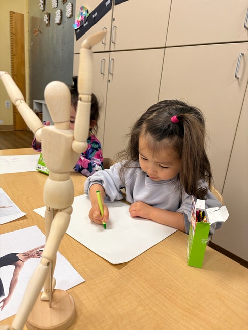A child drawing with crayons at a table beside a wooden mannequin used for art.