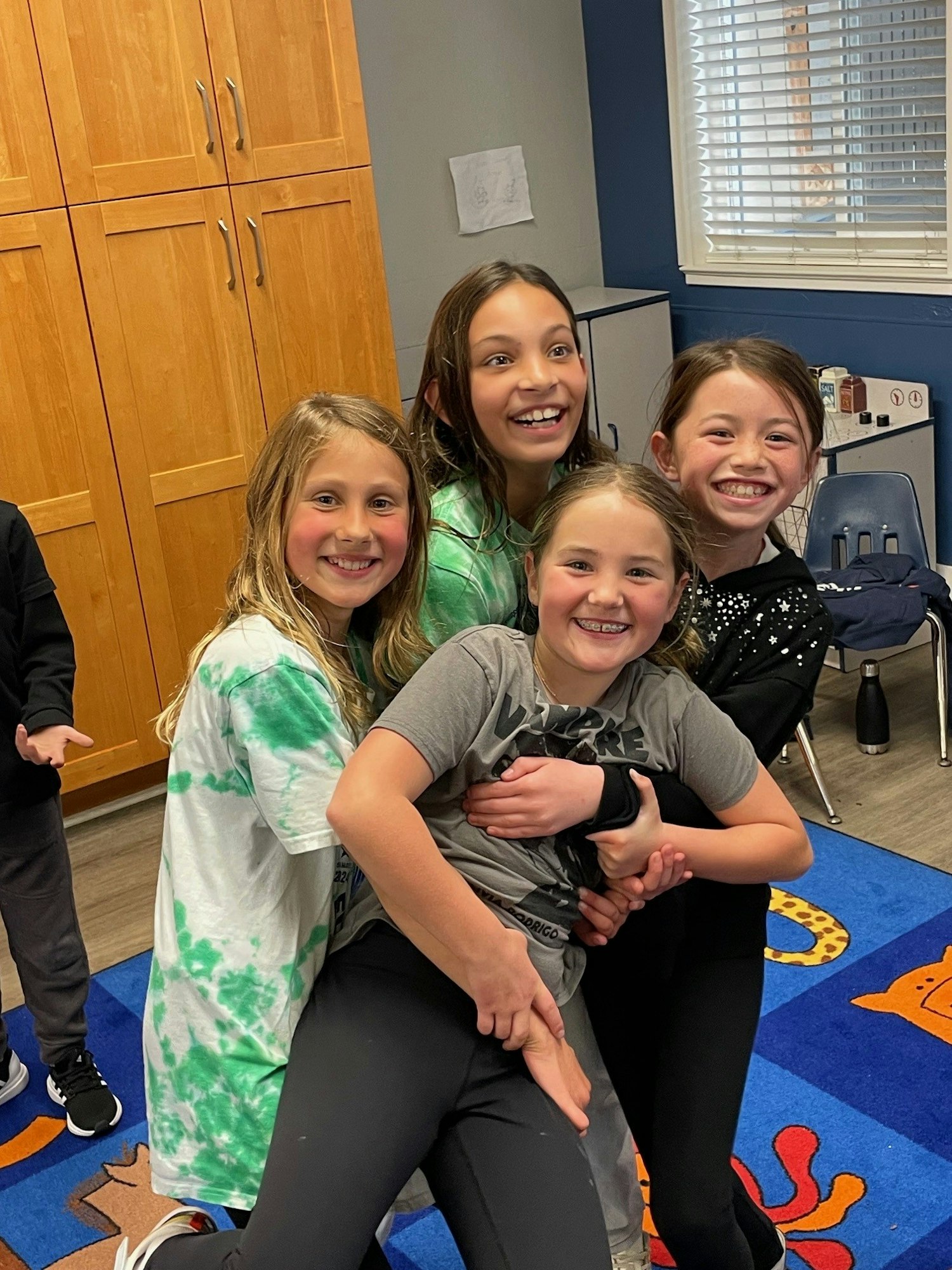 Four children smiling, three lifting another, in a room with cabinets and colorful carpet.
