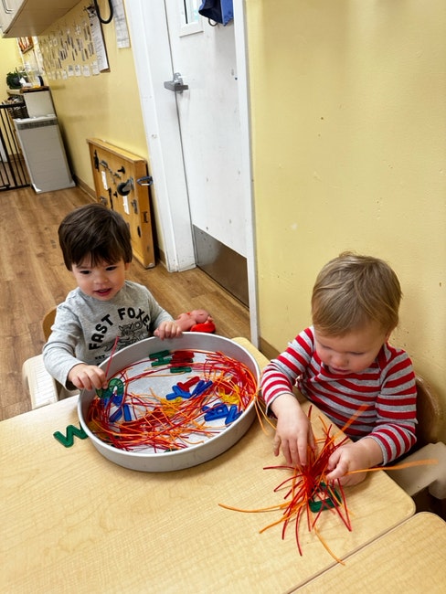Two toddlers playing with colorful strings and letters at a table.