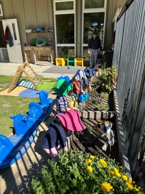 Children in hats playing and gardening outside with toys and tools in a sunny yard.