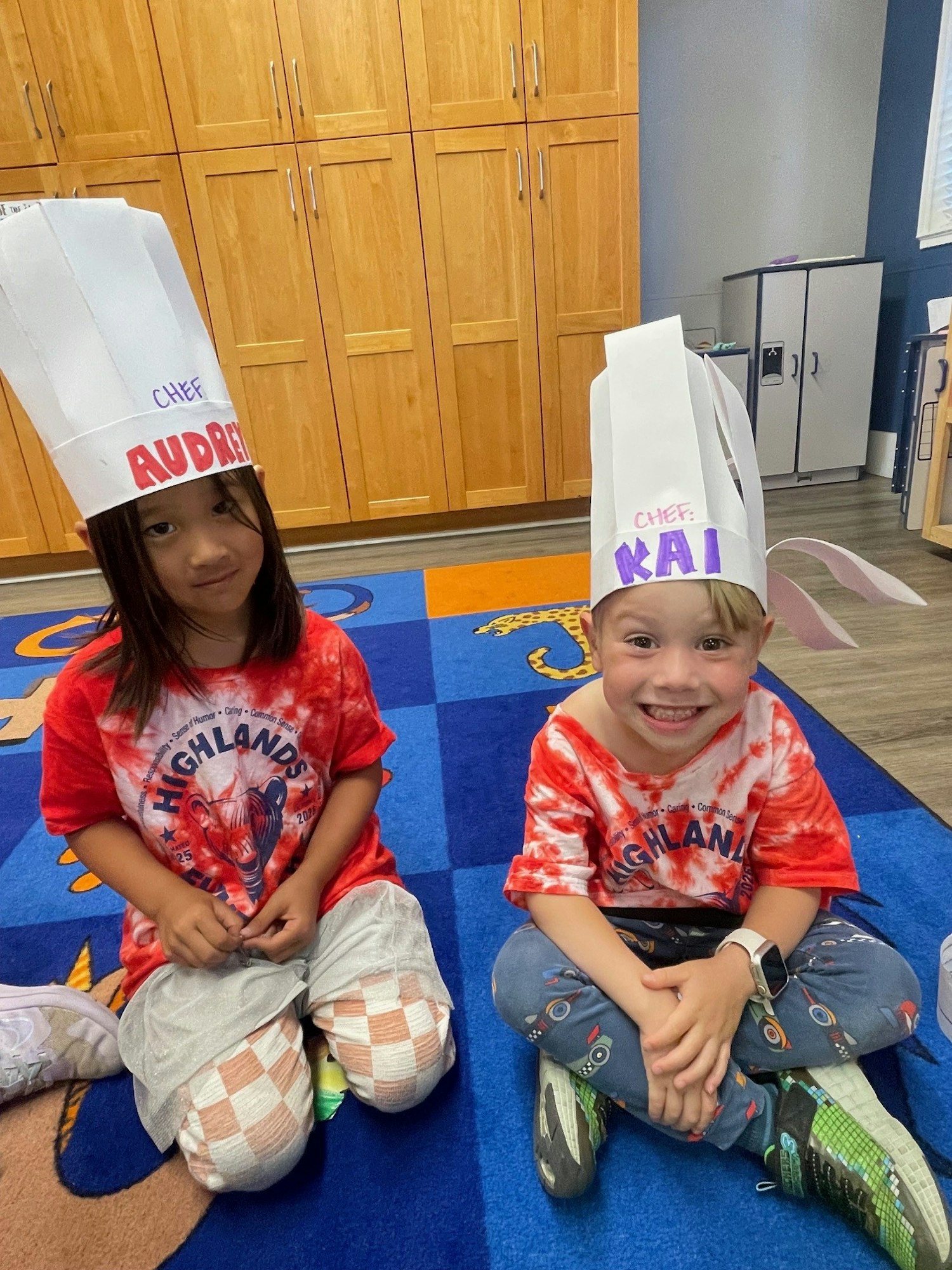 Two kids sitting on a colorful carpet wearing red tie-dye shirts and paper chef hats with names written on them.