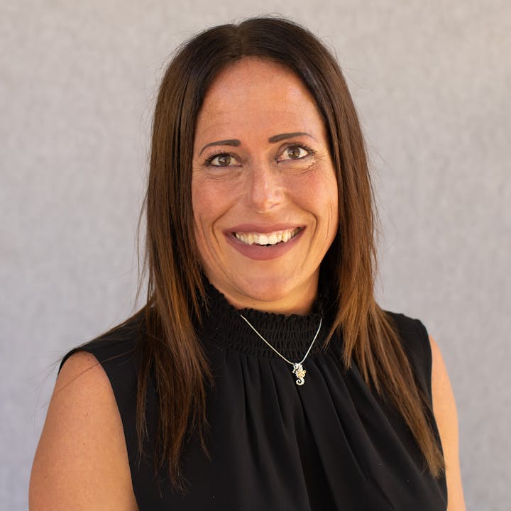 The image shows a smiling woman with long brown hair, wearing a black top and a necklace, against a light background.