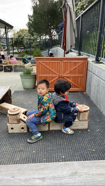 Two kids are sitting back-to-back on a wooden structure outdoors, surrounded by a playground area.