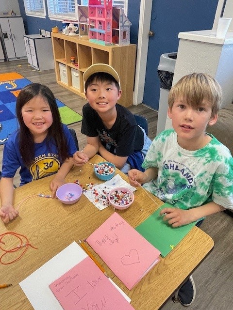 Three kids at a table with craft supplies and colorful papers, smiling and engaging in an activity.