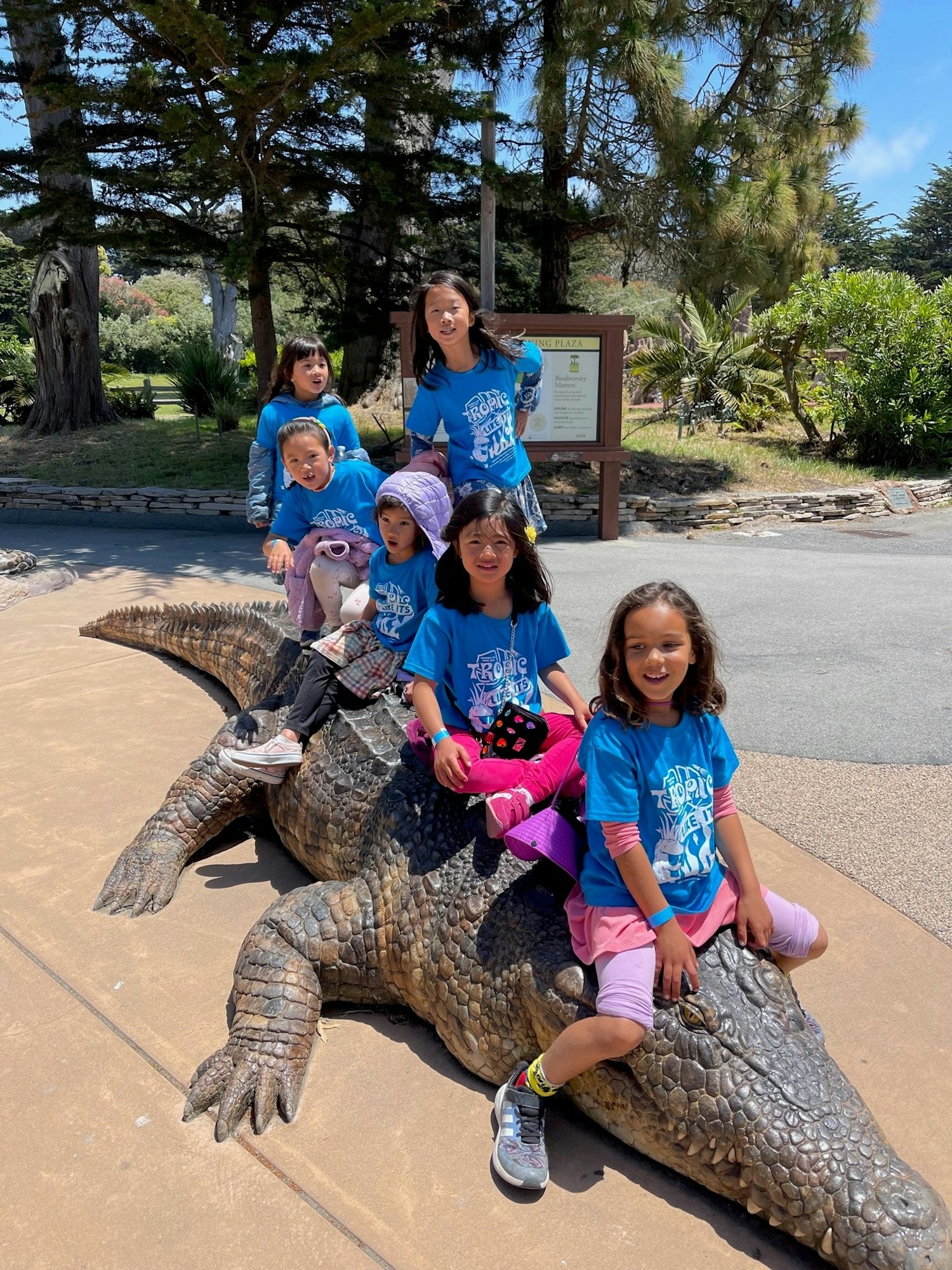A group of six children in blue shirts poses playfully on a large crocodile statue outdoors on a sunny day.