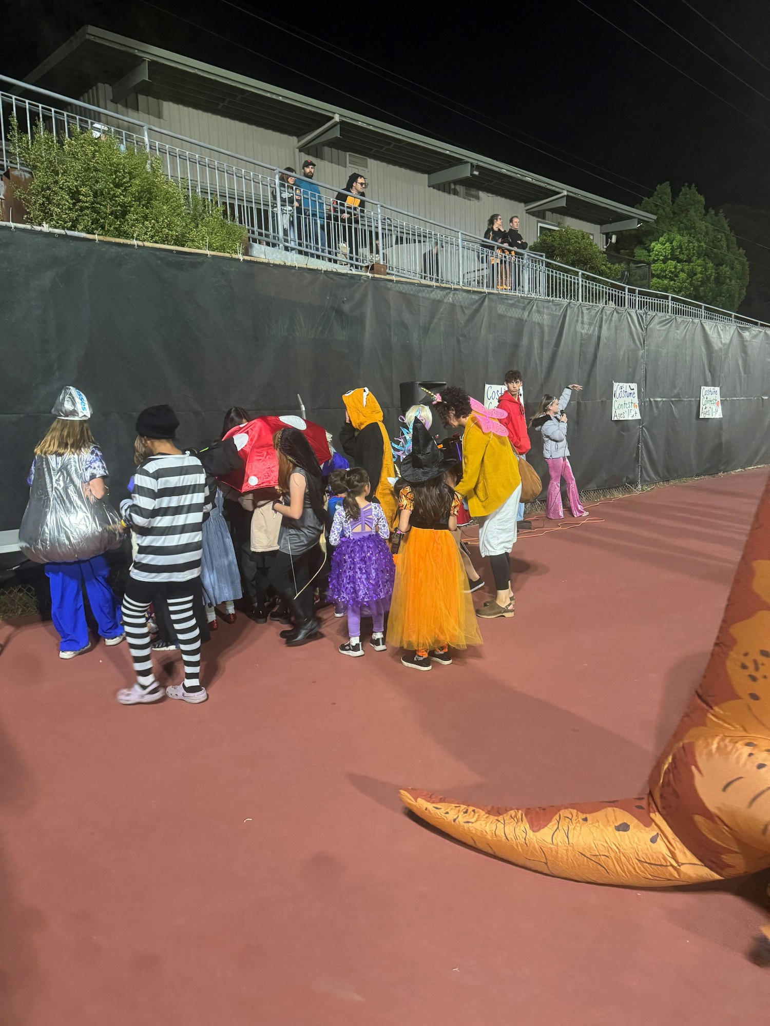 Children in costumes gather at an event, with onlookers above on a balcony. A large inflatable dinosaur tail is visible.