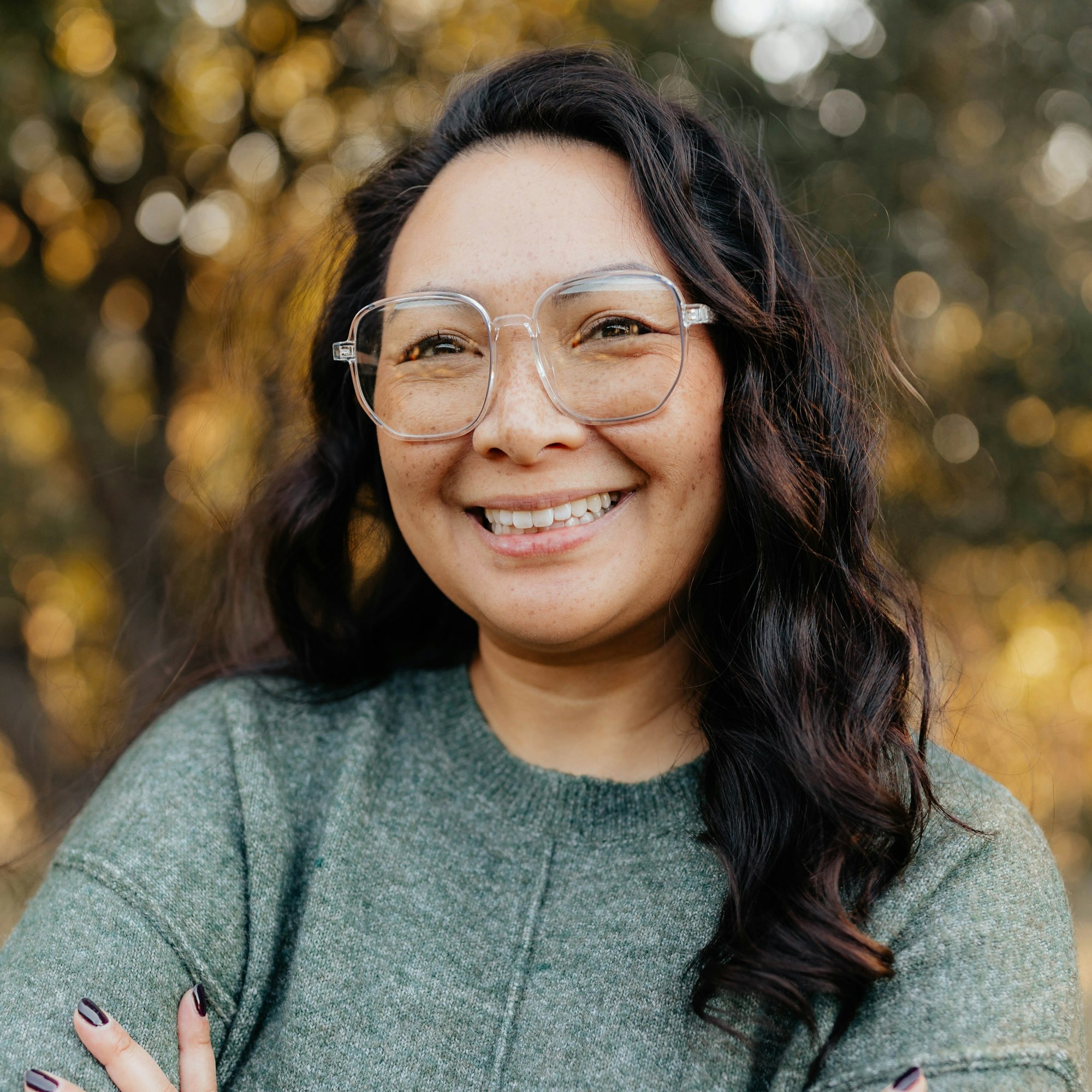 A person with glasses smiling, wearing a green sweater, standing outdoors with a blurred, leafy background.