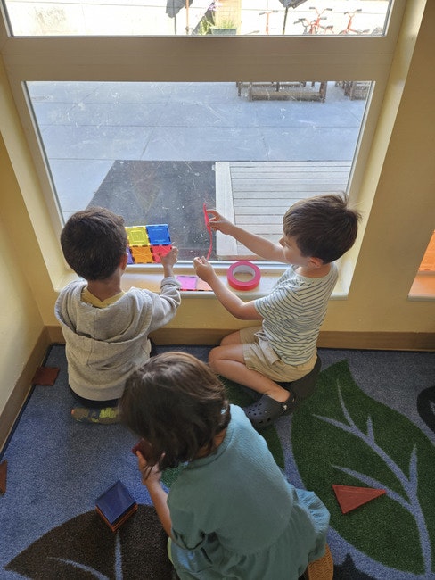 Three children playing with colorful blocks by a window.