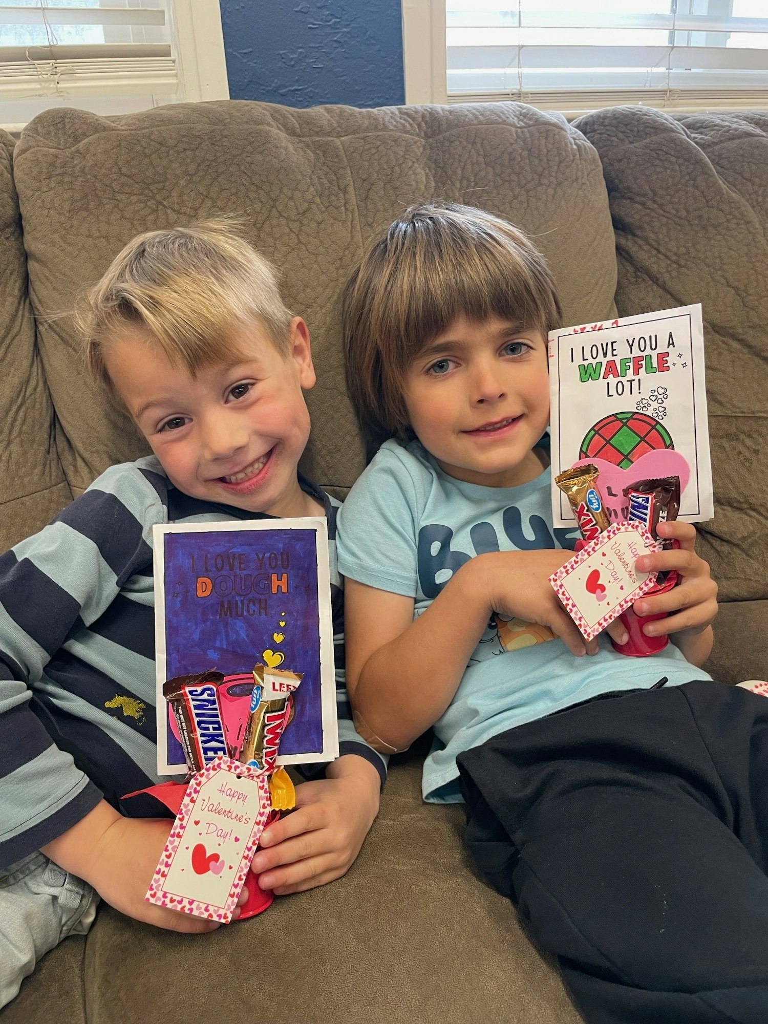 Two smiling children holding Valentine’s Day cards and candy, sitting on a couch, celebrating the holiday with playful messages.