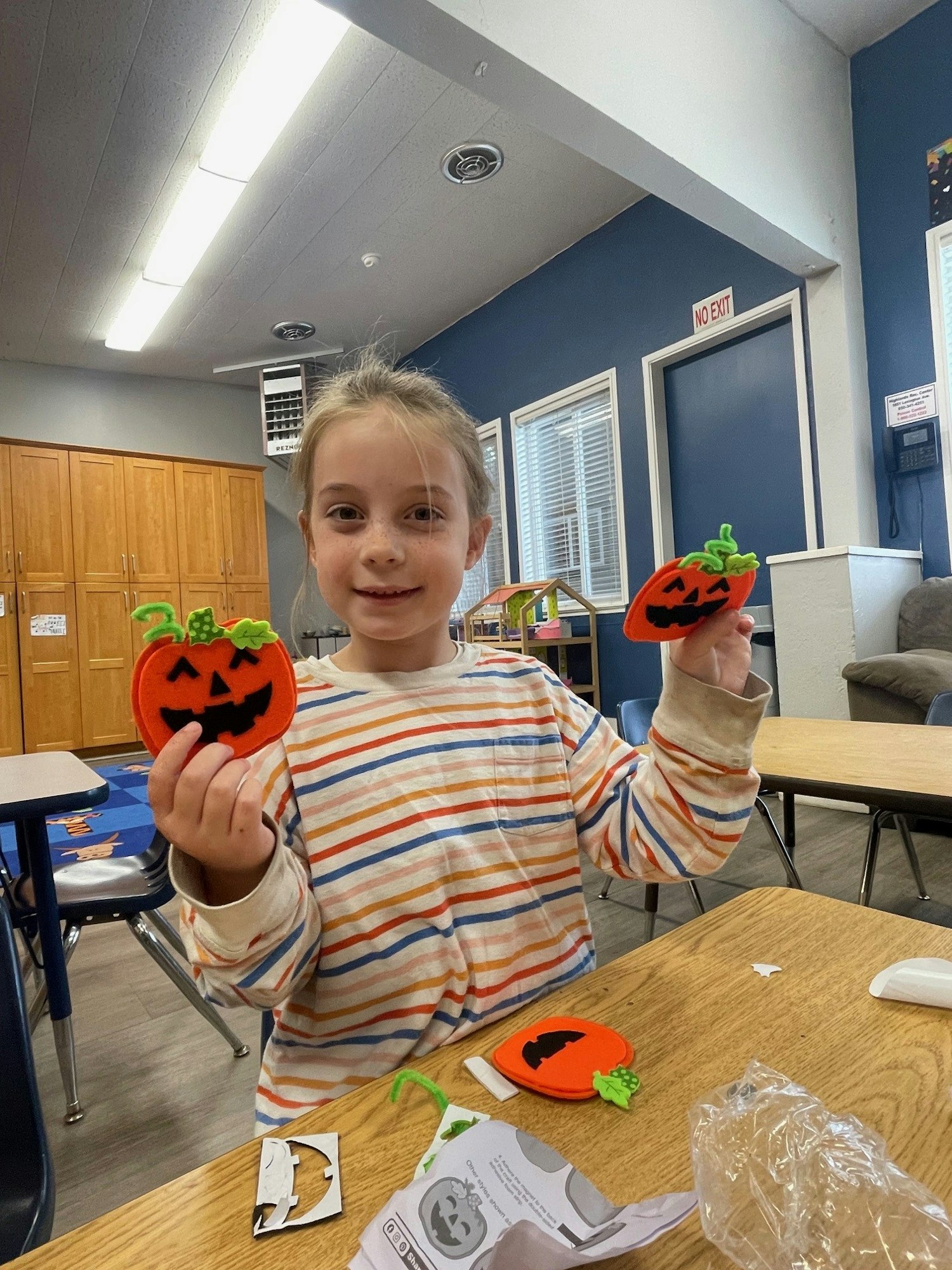 A child in a striped shirt holds two pumpkin crafts with black faces in a classroom setting.