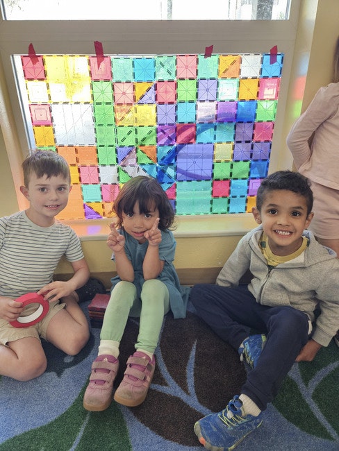 Three children sitting in front of a colorful, geometric stained glass-like window display.