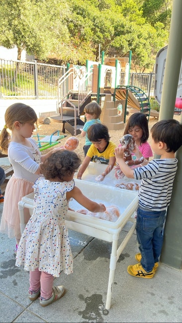 Children are playing with water and washing dolls in an outdoor area with playground equipment in the background.