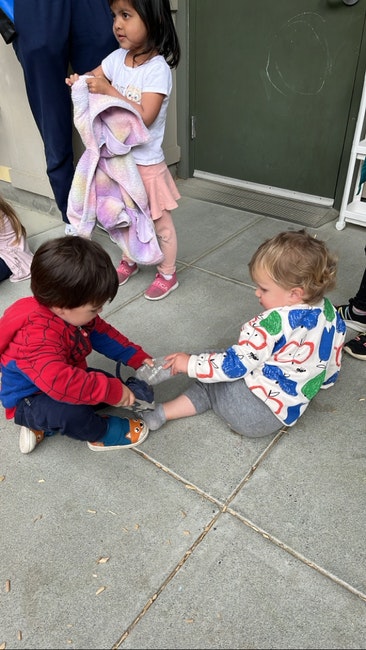 Three children are playing; one is sitting, another is helping, and a third is standing holding a colorful blanket.