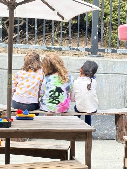 Three children sitting on a bench outdoors, seen from behind, facing a fenced area.