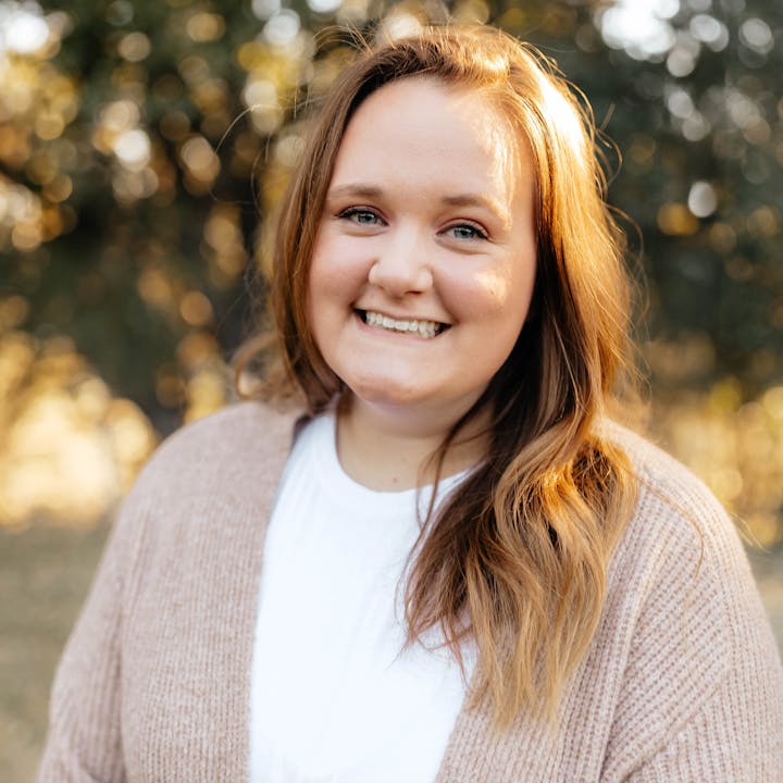 A person smiling outdoors with a blurred background of trees.
