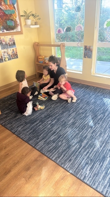 A group of children and an adult sit on a blue rug with books in a bright room with windows and artwork on the walls.