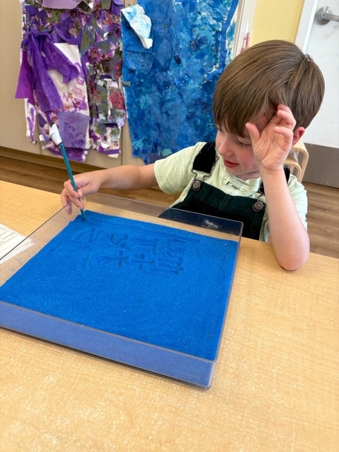 A child draws in a tray filled with blue sand using a pencil, sitting at a table. Art is displayed in the background.