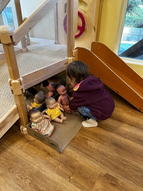 A child plays with several baby dolls under a wooden structure near a small slide indoors.