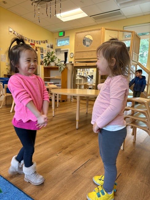 Two young children smiling at each other in a classroom setting with wooden furniture and decorations.