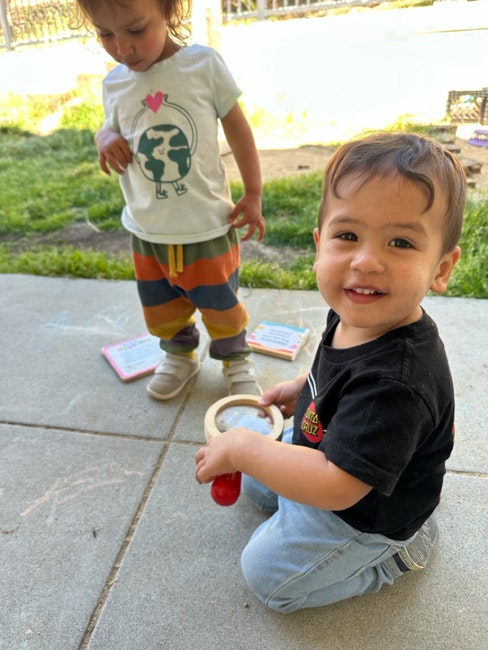 Two children are playing outside on a patio. One is holding a toy drum, and the other is standing nearby with colorful pants.