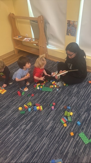 Children playing with building blocks on the floor while an adult reads to them.