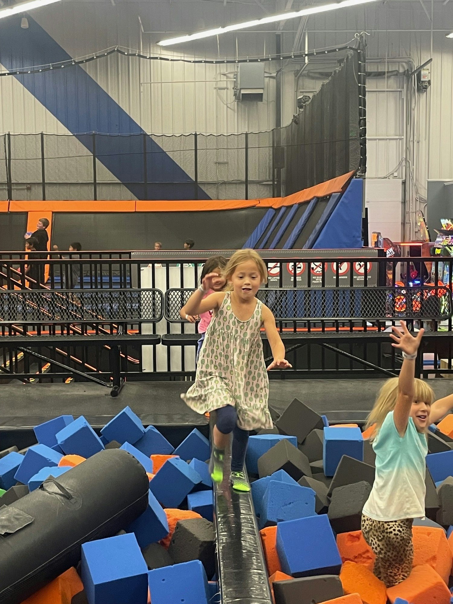 Children playing on a balance beam over a foam pit in an indoor playground with trampolines in the background.