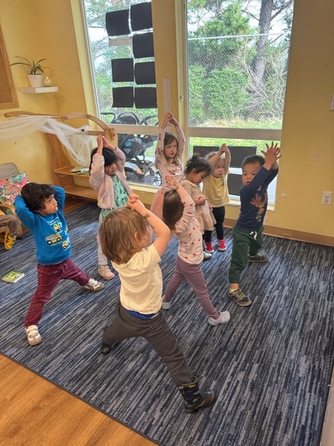 Children in a classroom doing yoga poses with hands raised.