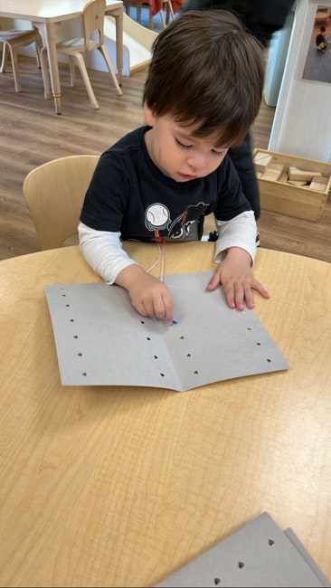 A child is threading a colorful string through holes in a piece of paper, seated at a wooden table in a classroom setting.