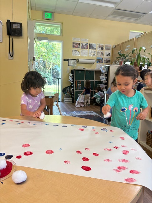 Children painting with colored dots on a large sheet of paper in a classroom.
