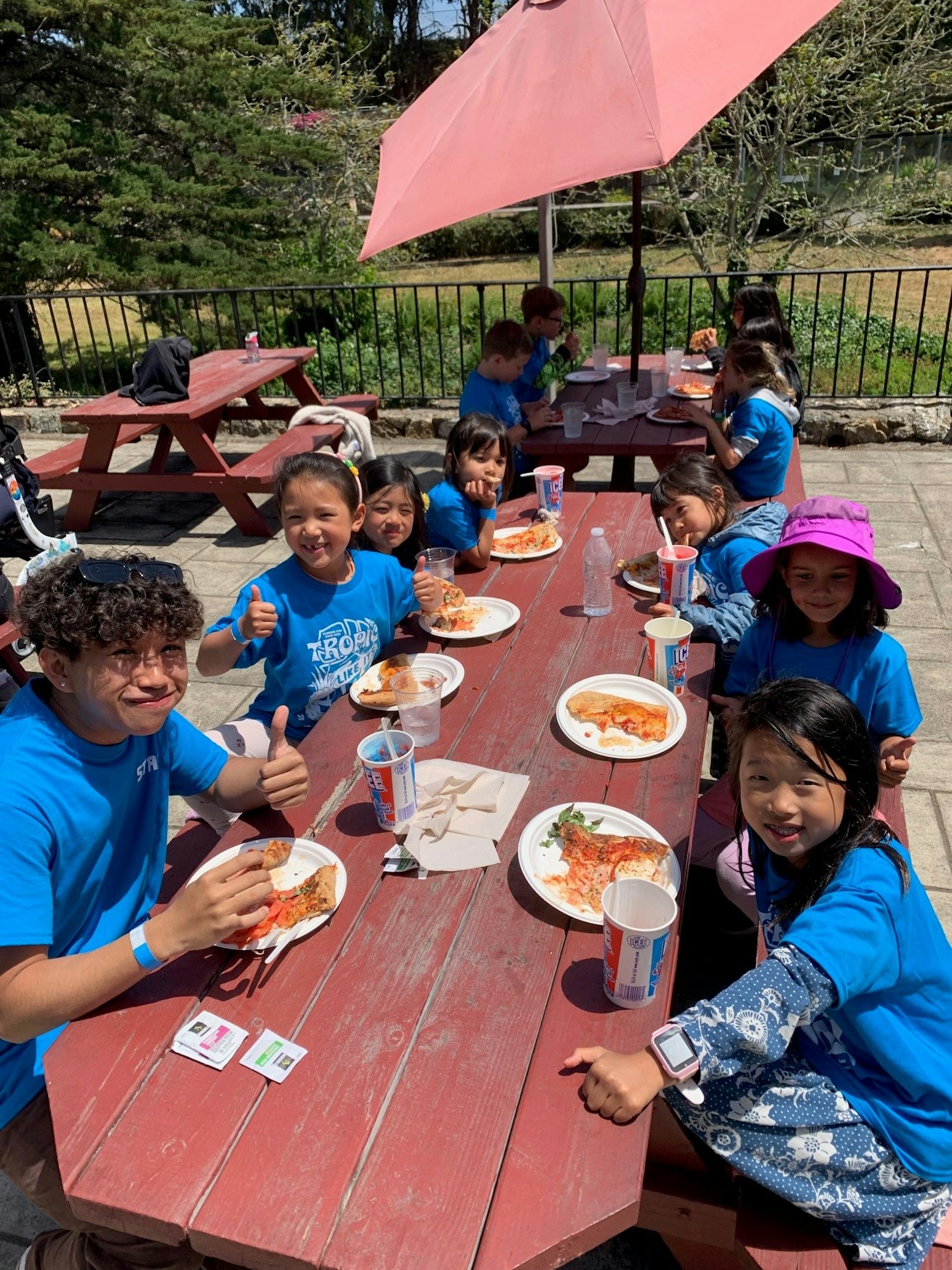 A group of children in blue shirts enjoying pizza and drinks at an outdoor picnic table on a sunny day.