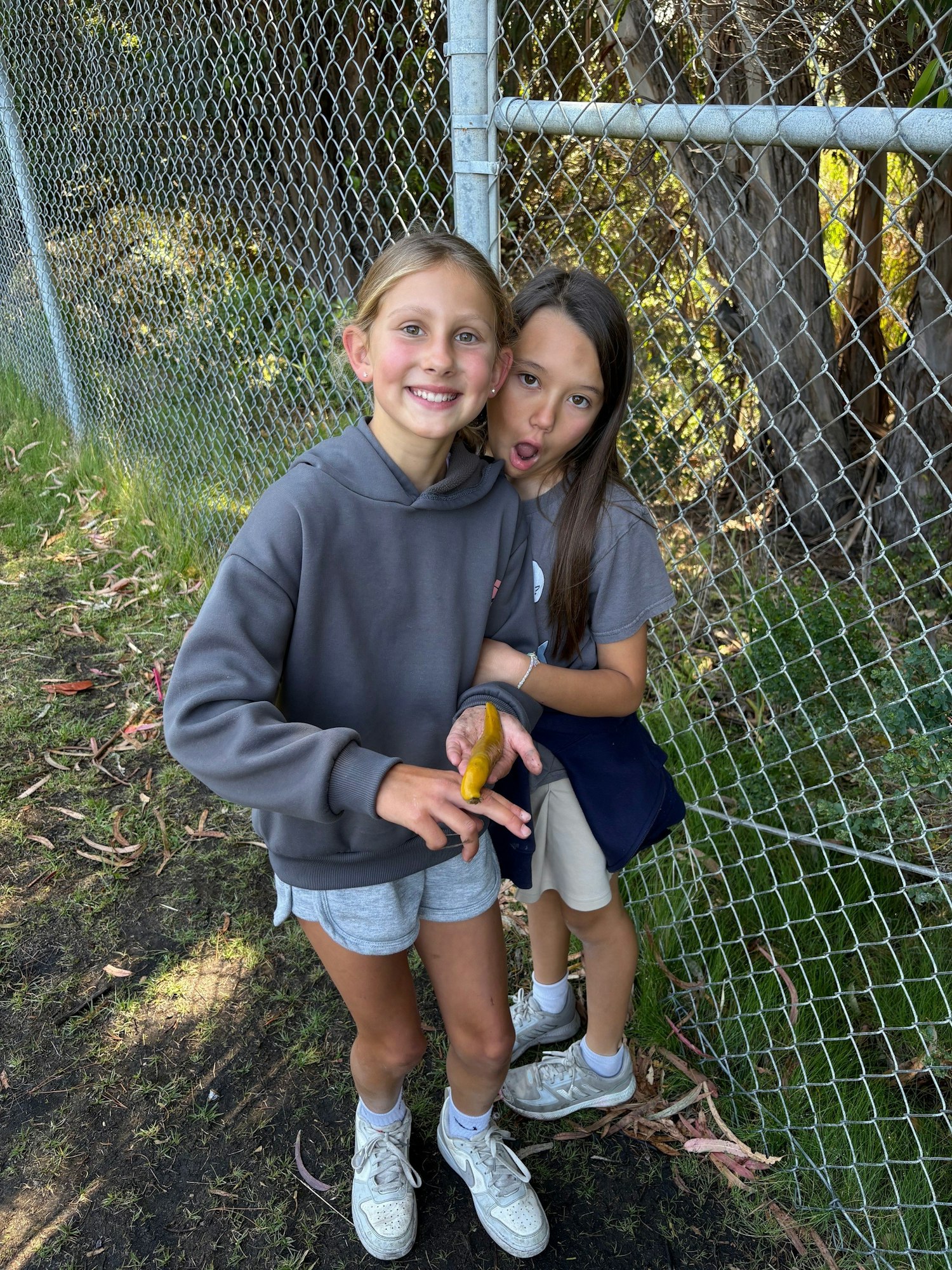Two kids smiling by a chain-link fence. One is holding a banana slug on her finger.