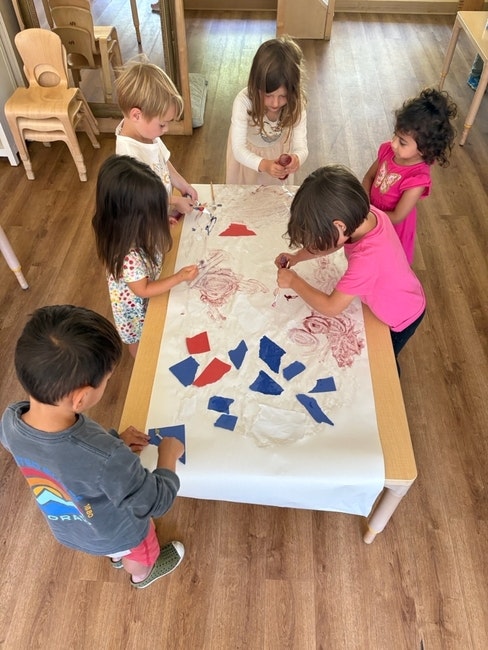 Children are gathered around a table doing a collaborative art project with red and blue paper pieces.