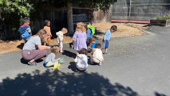 Adults and children gathered outdoors, some squatting, with trees in the background.
