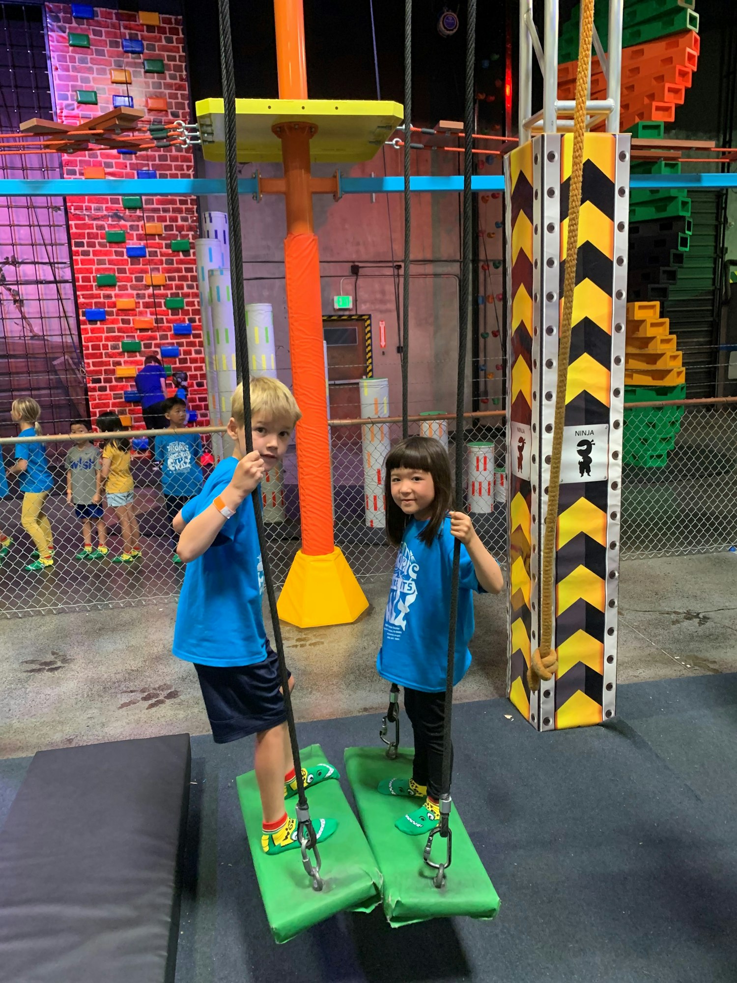 Two kids on swings in a colorful indoor play area, surrounded by climbing structures and a lively atmosphere.