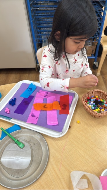 Child crafting with colorful papers and beads on a tray; basket of pom-poms nearby.