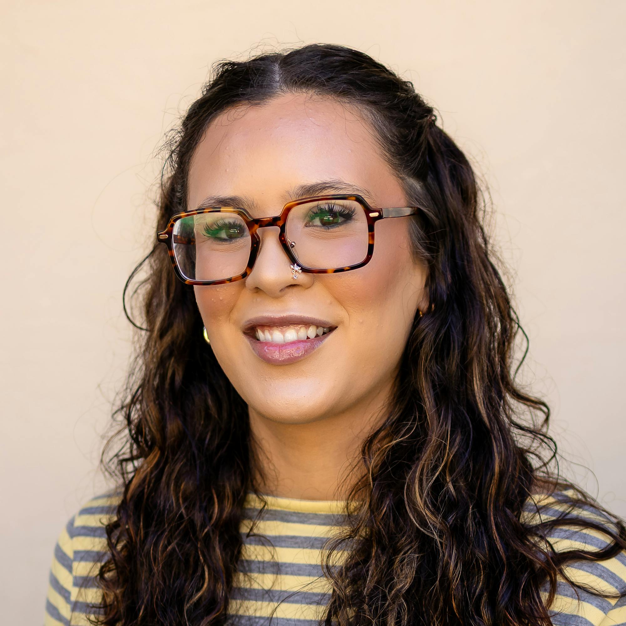 A woman with curly hair and stylish eyeglasses smiles, wearing a striped long-sleeve shirt against a neutral background.