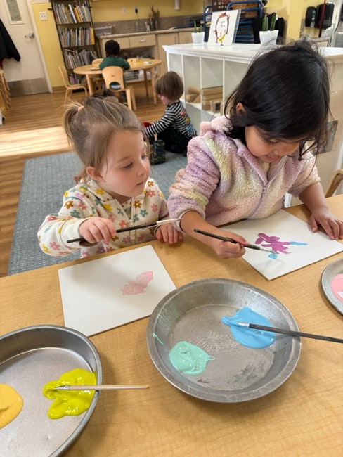 Two children painting with brushes at a table, using colorful paints in pans.