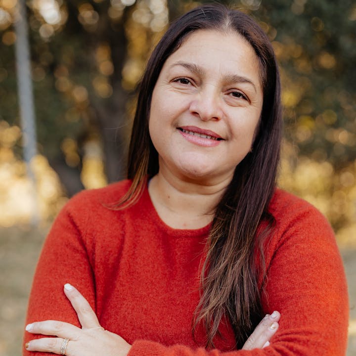 A person wearing a red sweater with arms crossed, smiling at the camera. Background features blurred greenery.