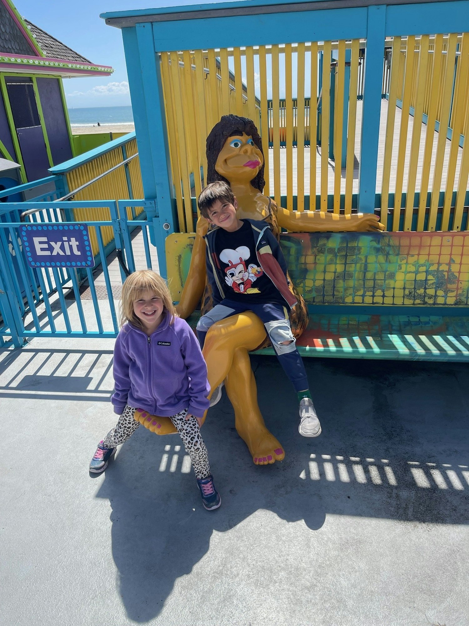 Two kids smiling near a yellow statue on a boardwalk, with an "Exit" sign and ocean in the background.